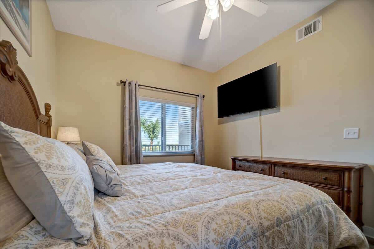 Un dormitorio con cama, televisor montado en la pared, ventana con cortinas y ventilador de techo.