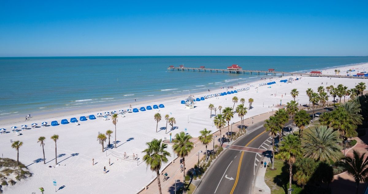 Pristine white sands meet crystal-clear turquoise waters along this stunning Gulf Coast beach, framed by swaying palms and a historic fishing pier extending into the calm sea.
