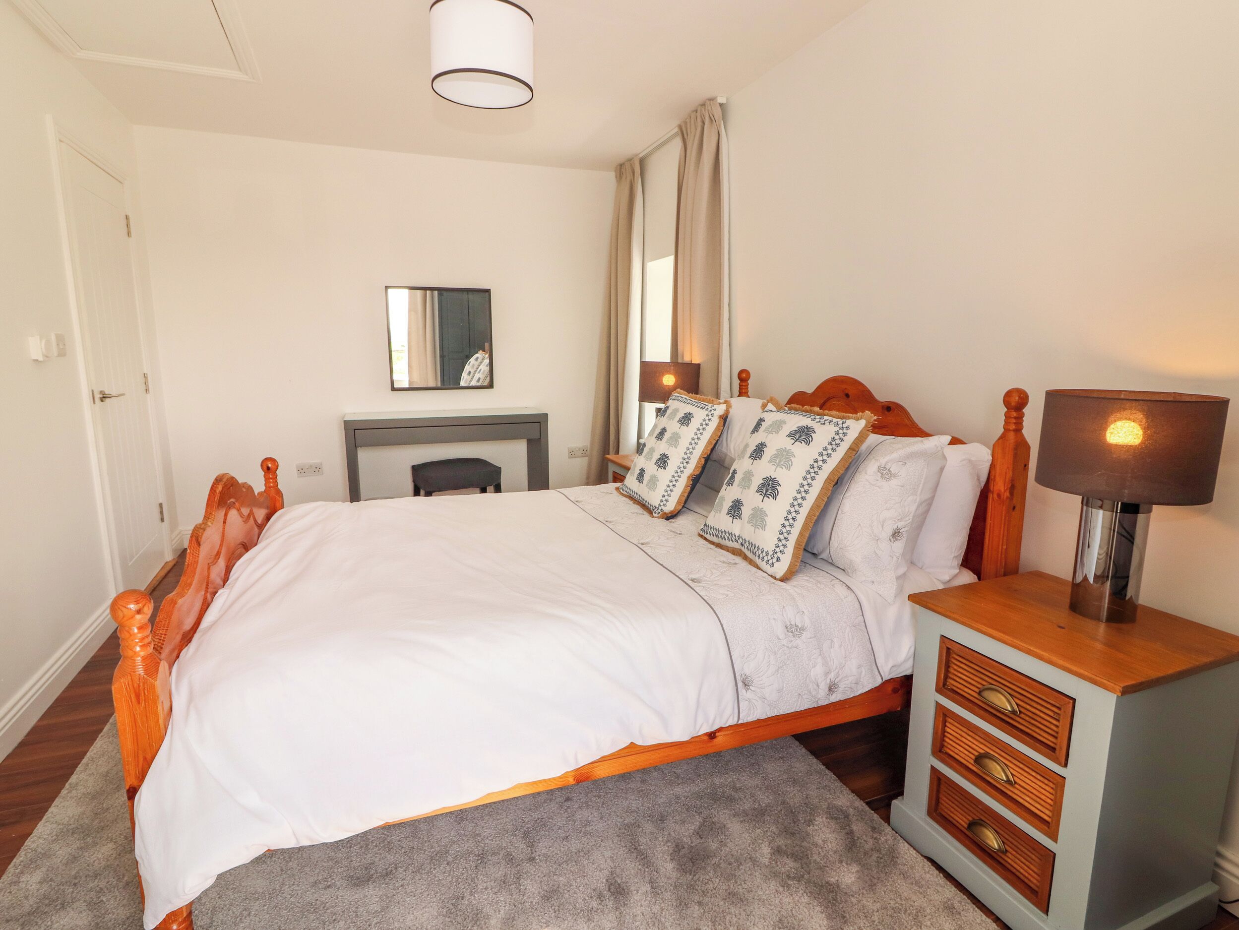 A neatly arranged bedroom with a bed, bedside tables, a lamp, a mirror, and a small seating area.
