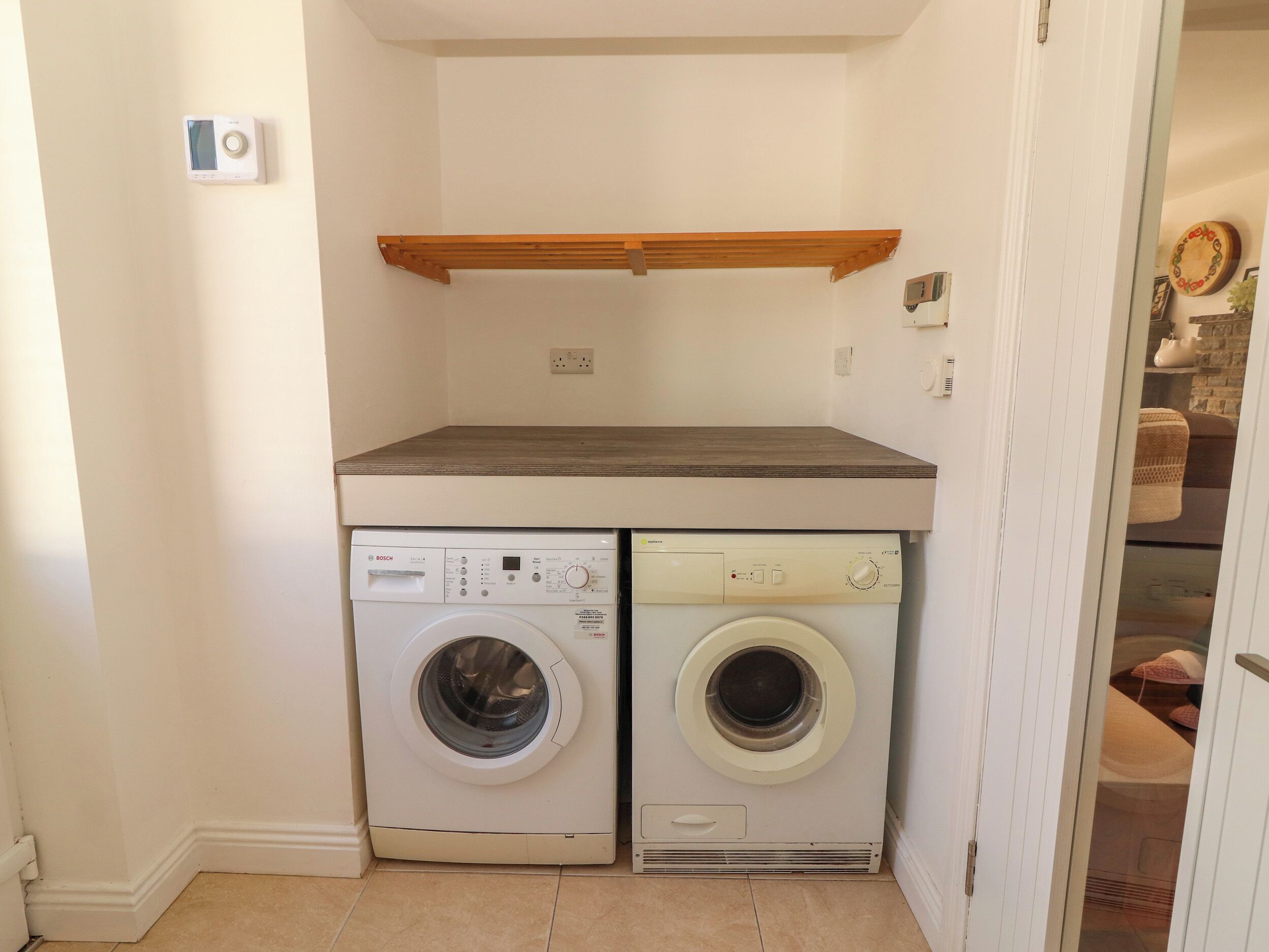 A laundry room with two front-loading washing machines, a wooden shelf above, and a tiled floor.