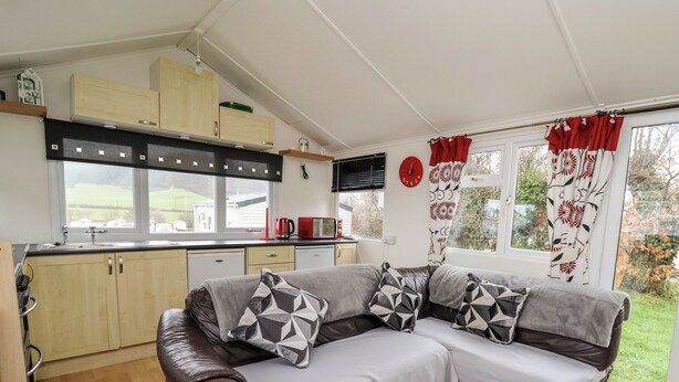 A cozy living area with a sofa, a kitchen with wooden cabinets, a window with curtains, and a view of greenery outside. - Chalet 32 (Aberystwyth)