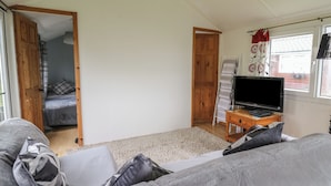 A room with a grey sofa, a TV on a wooden stand, a wardrobe, and a door leading to another room. - Chalet 32 (Aberystwyth)