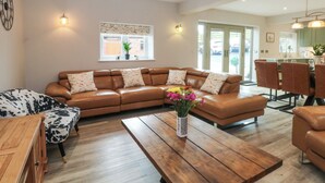 A modern living room with a brown leather sectional sofa, a wooden coffee table with a vase of flowers, and a dining area with a table and chairs. - Small Lane Barn (Stoke-on-Trent)