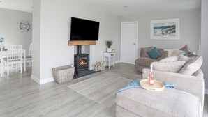 Ein modernes Wohnzimmer mit Kamin, einem flachbildfernseher darüber, einer Couch, einem Essbereich mit Stühlen und einem kleinen Tisch mit einer Pflanze.