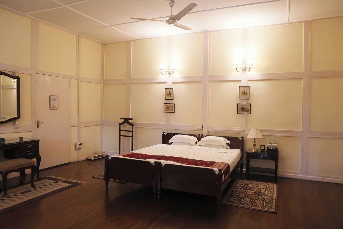 Quarto de hotel com cama de madeira, mesa de cabeceira com abajur, um banco, escrivaninha e espelho.