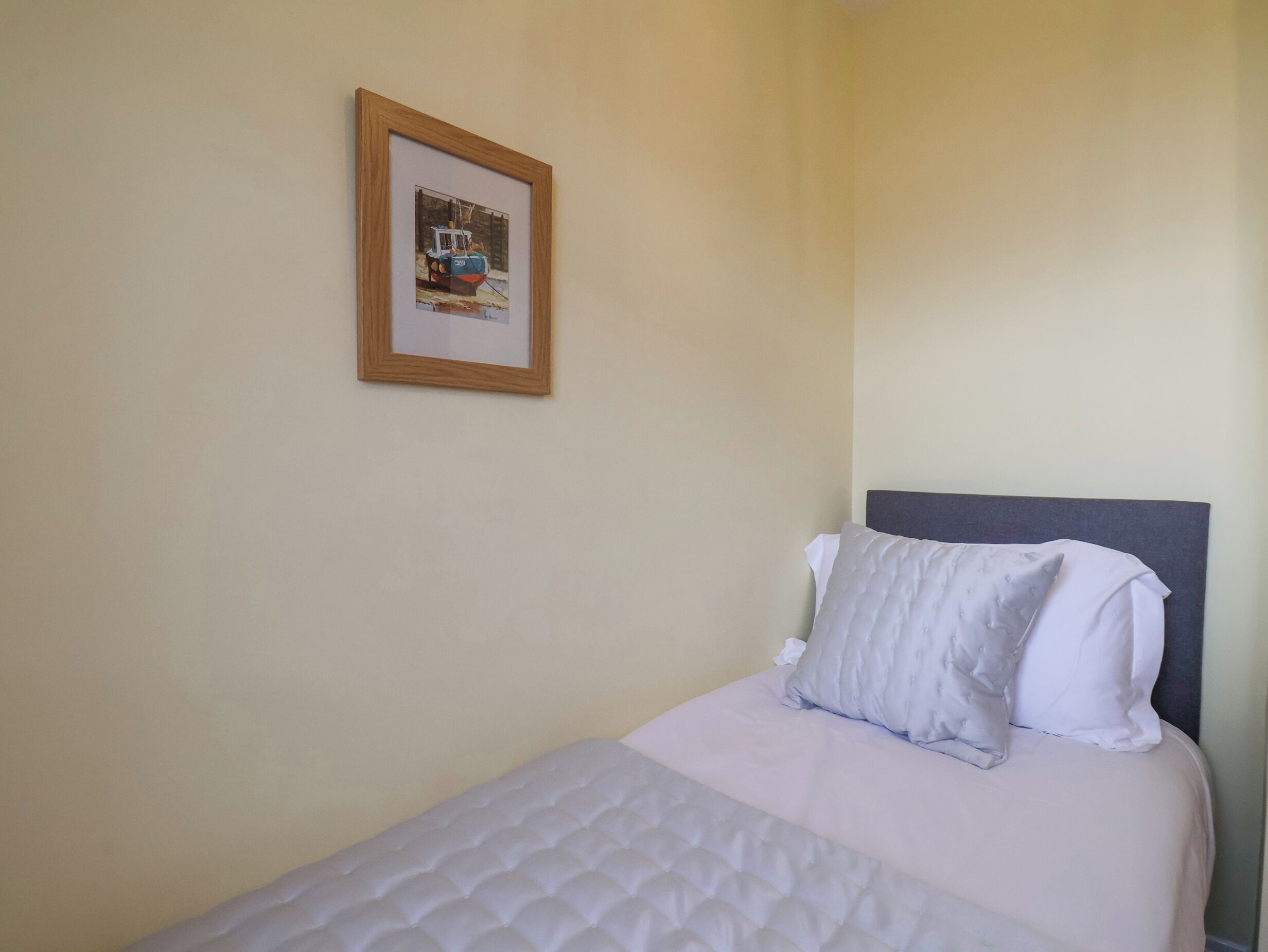 A single bed with a white quilted bedspread and two pillows, a framed picture on the wall, and a plain wall.