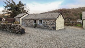 A stone cottage with a gravel driveway, a wooden fence, and a large tree in the background.