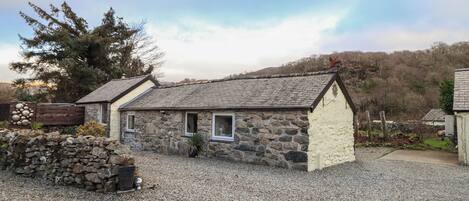 A stone cottage with a gravel driveway, a wooden fence, and a large tree in the background.
