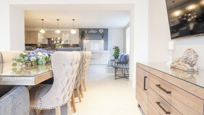 Una cocina moderna con comedor, que incluye una mesa de vidrio, sillas tapizadas, iluminación colgante y un televisor de pantalla plana montado en la pared.