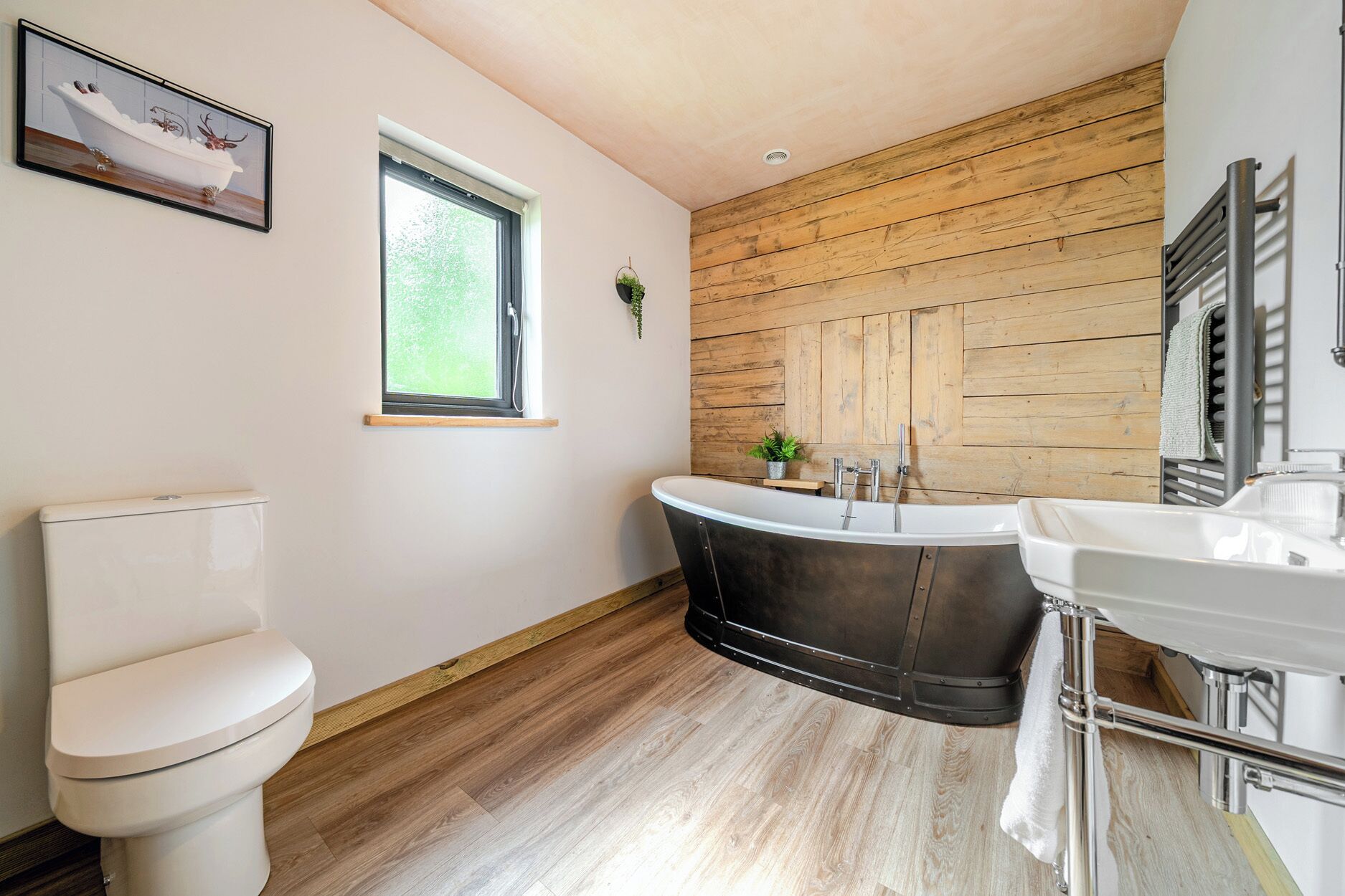 Un baño con inodoro, bañera independiente, lavamanos, ventana y pared de madera.