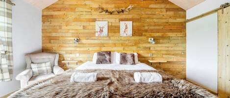 A bedroom with a wooden slat ceiling, a bed with a fur throw, a chair, and two wall-mounted lamps.