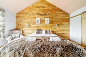 A bedroom with a wooden slat ceiling, a bed with a fur throw, a chair, and two wall-mounted lamps.