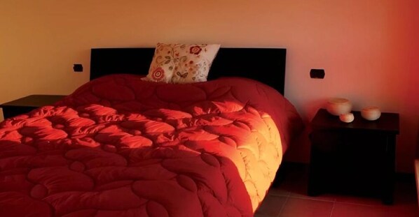 A bedroom with a large bed, a nightstand with a lamp, and a wall-mounted shelf with decorative items. - Agriturismo Prisma: Quadruple Room with Private Bathroom (Villa Vergano)