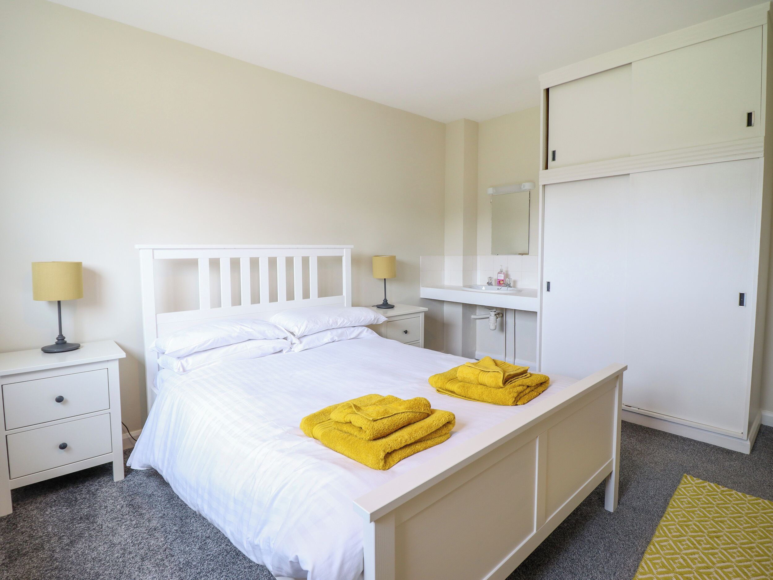 A neatly arranged bedroom with a bed, two bedside tables, a nightstand, a wardrobe, and a bathroom with a sink and mirror.