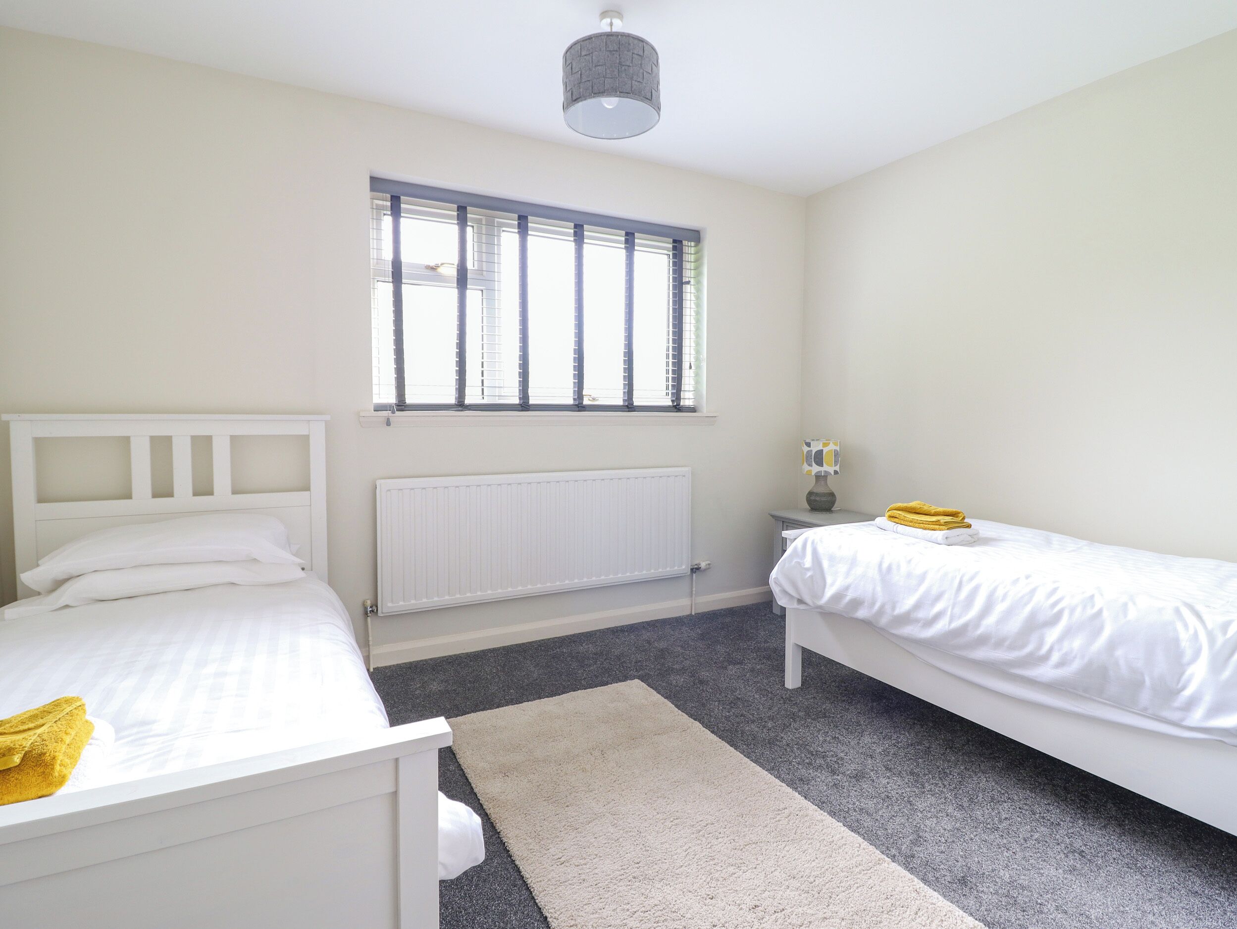A bedroom with two single beds, a radiator, a window with blinds, a lamp, and a small table.