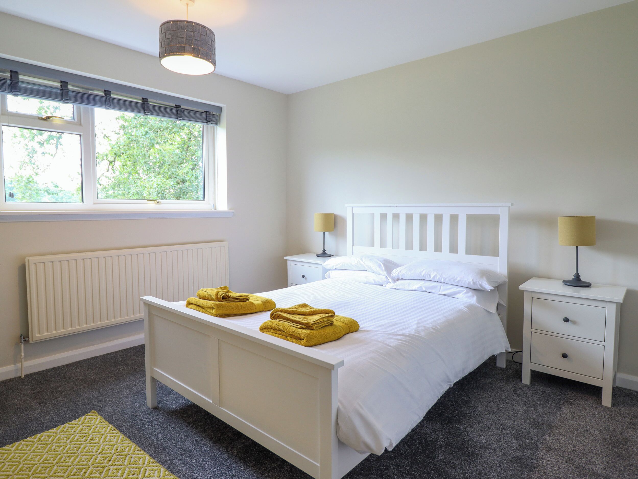 Une chambre à coucher avec un lit blanc, deux tables de chevet avec des lampes, un radiateur et une fenêtre avec des stores.
