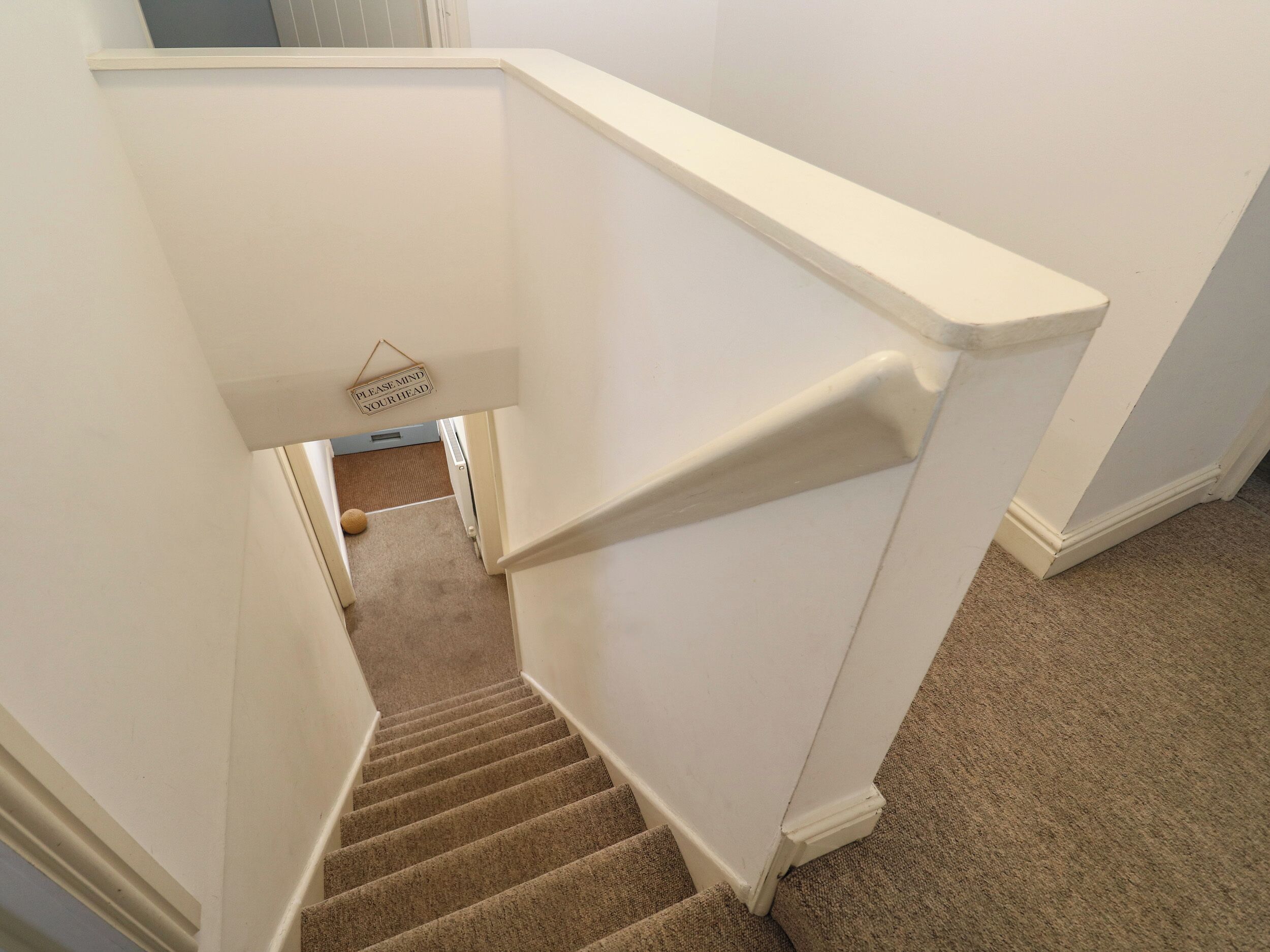 A carpeted staircase with white walls and a 'Please mind your head' sign at the top.