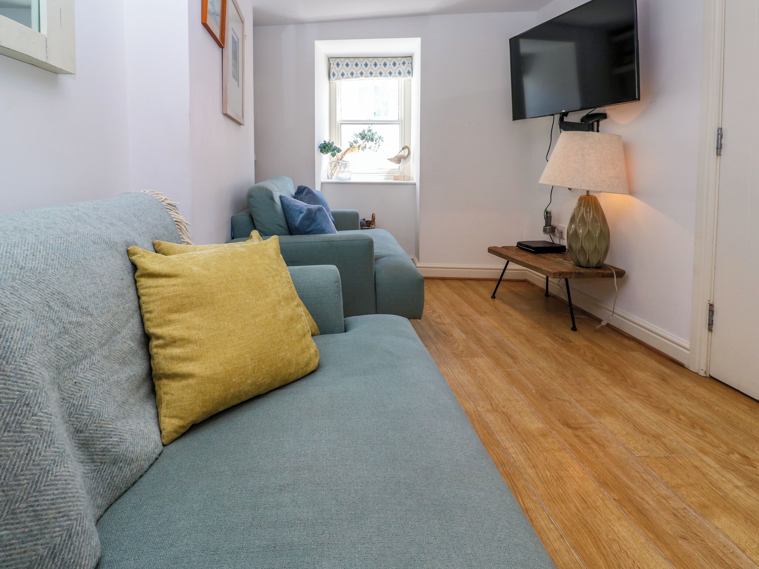 A living room with a grey sofa, a yellow pillow, a blue sofa, a wooden coffee table, a lamp, and a TV mounted on the wall.
