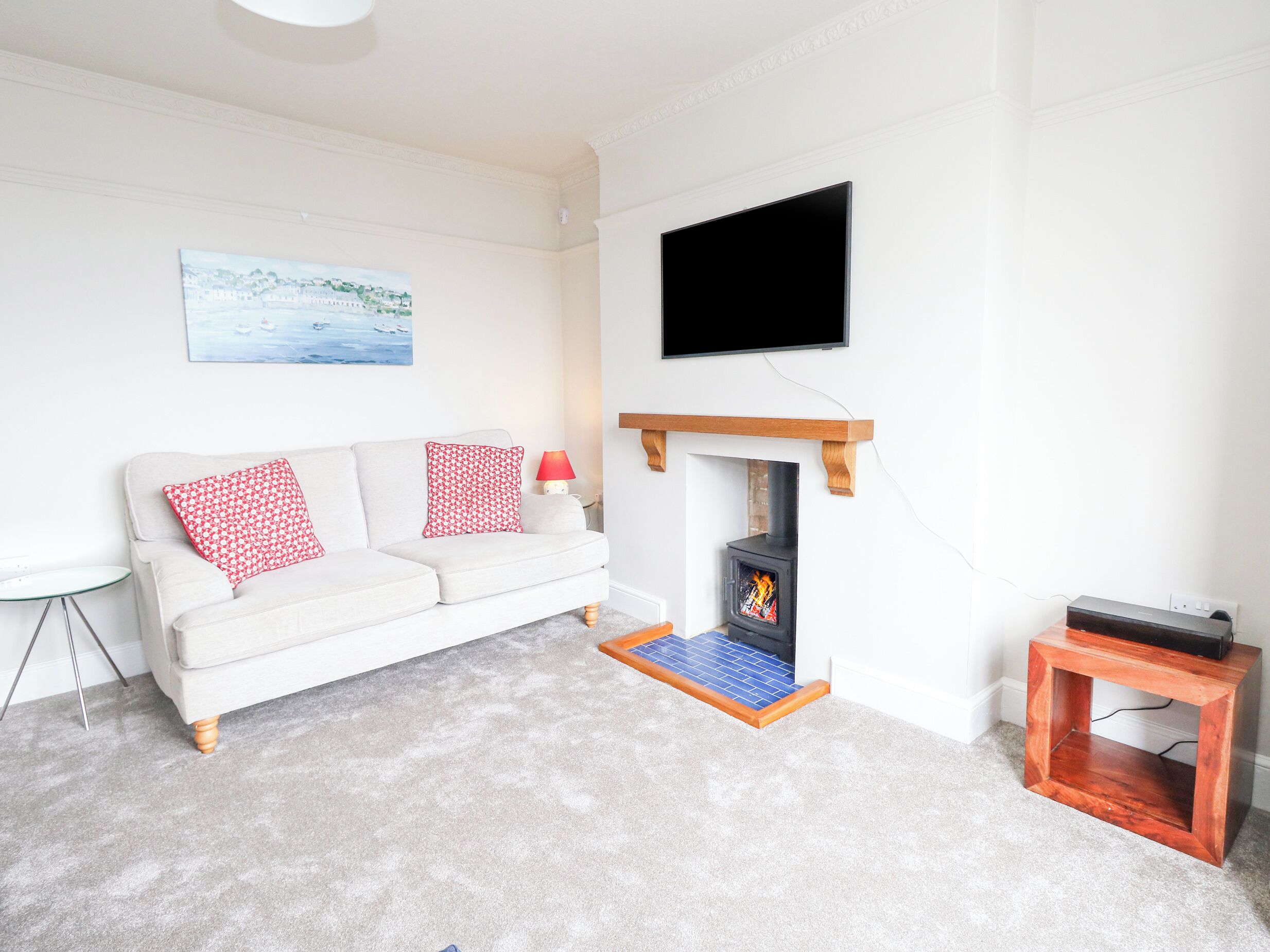 A living room with a white sofa, red and white patterned pillows, a flat-screen TV mounted above a fireplace, and a small wooden side table.