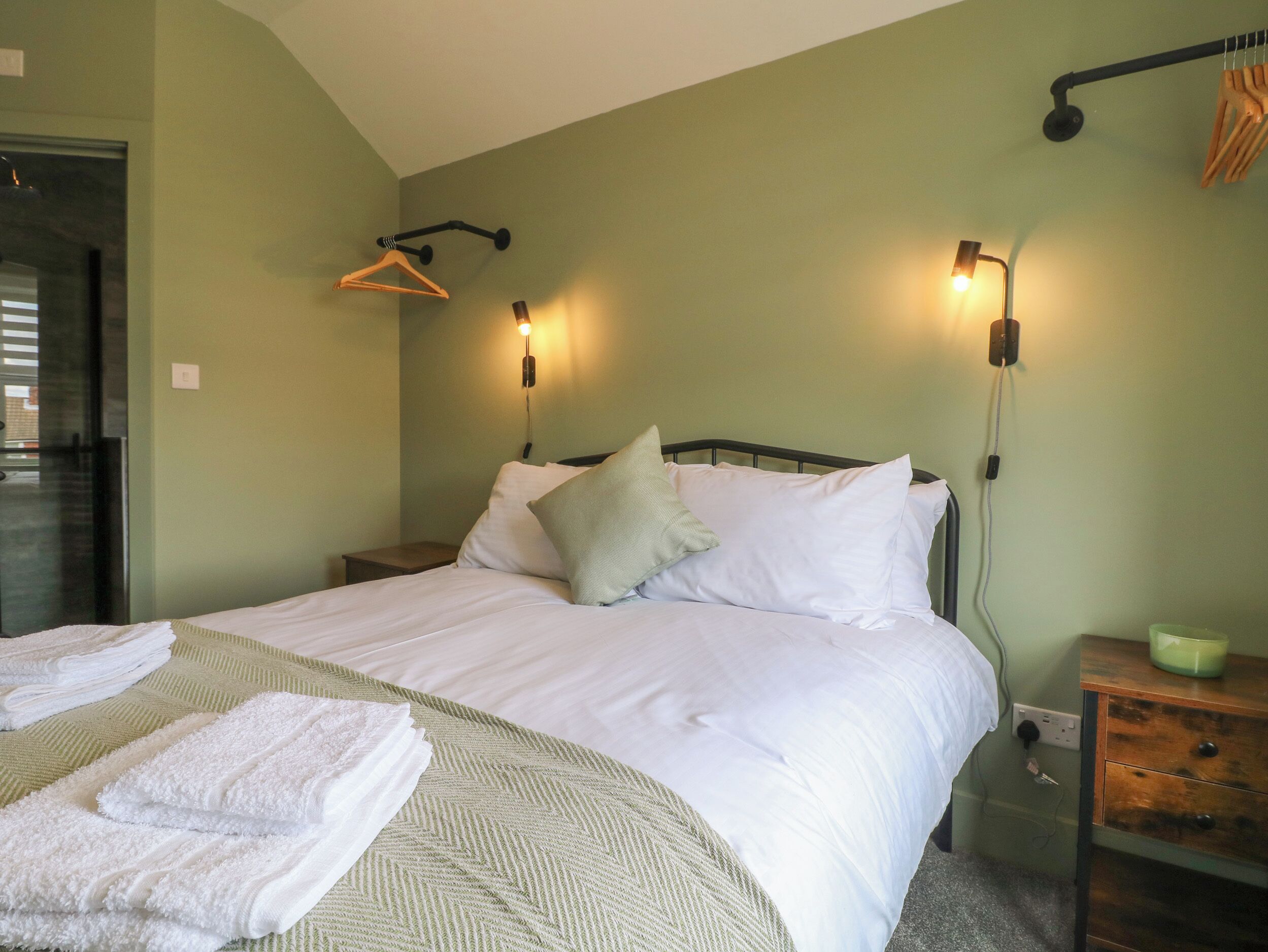 A neatly made bed with white linens and a green textured blanket, a bedside table with a lamp, and a wall-mounted light fixture.