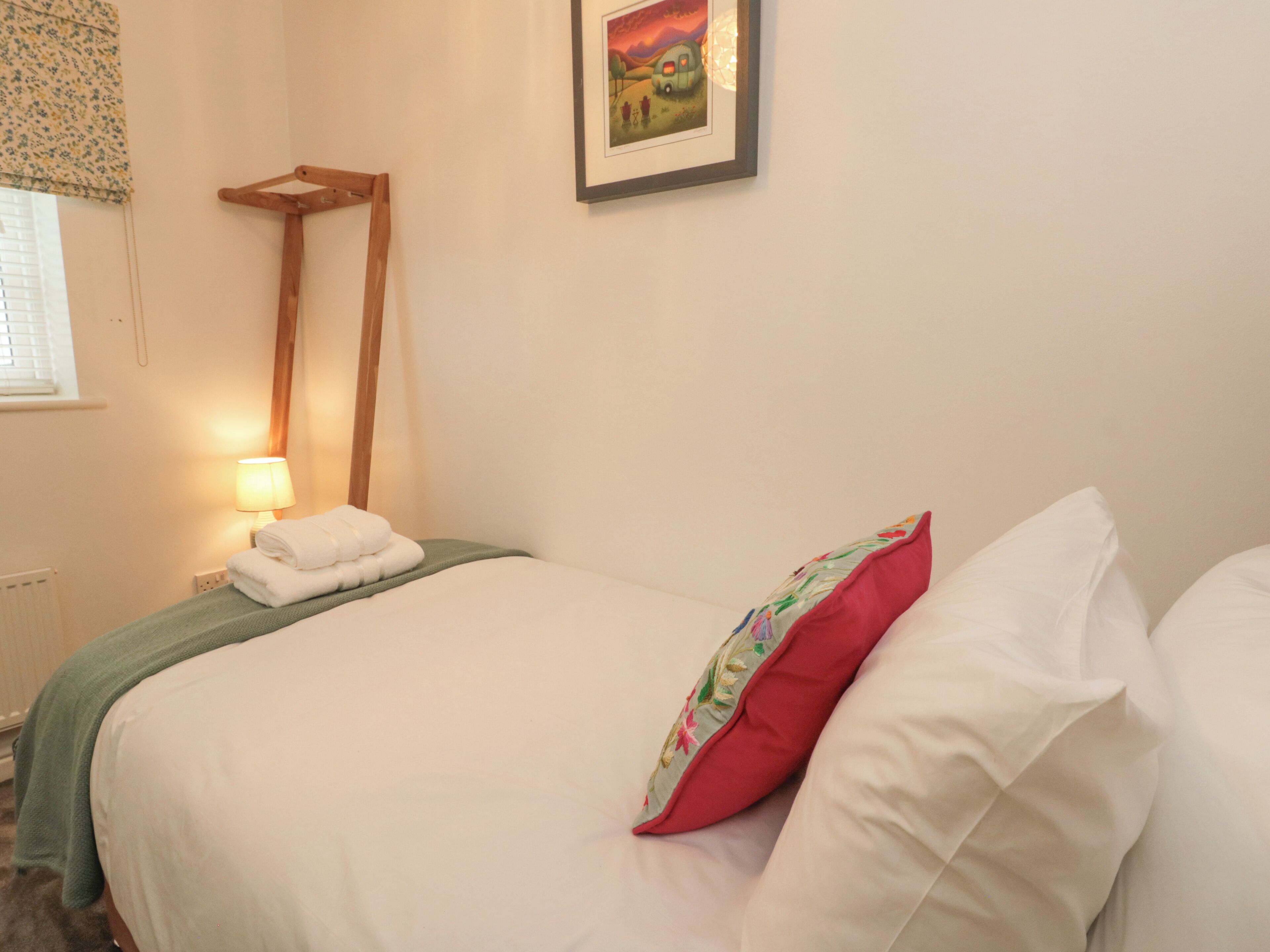 Una cama bien hecha con sábanas blancas y una manta verde, una mesita de noche de madera con una lámpara y un cuadro en la pared.