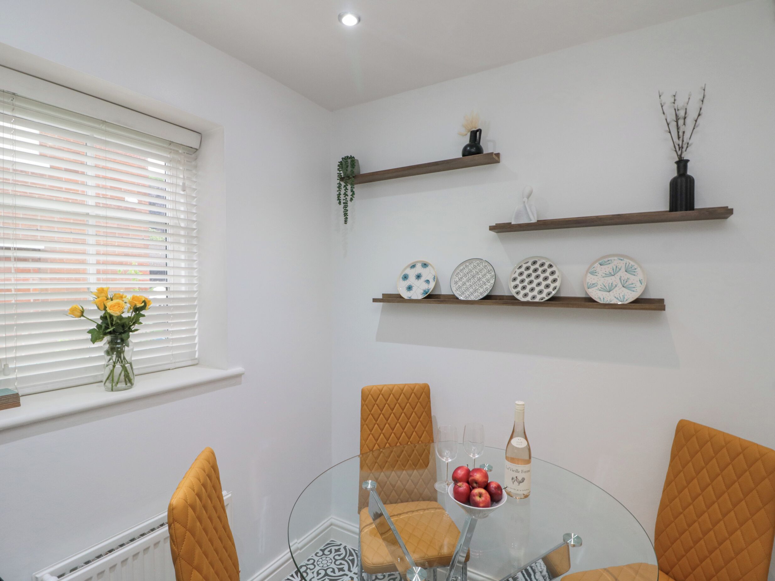 A dining area with a glass table, four chairs, a vase with yellow flowers, and decorative plates on shelves.