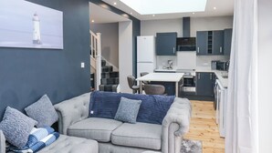 Een moderne woonkamer met een grijze bank, een eettafel en een keuken met donkere kasten en witte apparatuur.