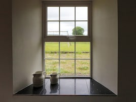 Una ventana con vistas a un campo cubierto de césped y un árbol. Debajo de la ventana, hay dos jarrones de cerámica sobre una superficie negra.