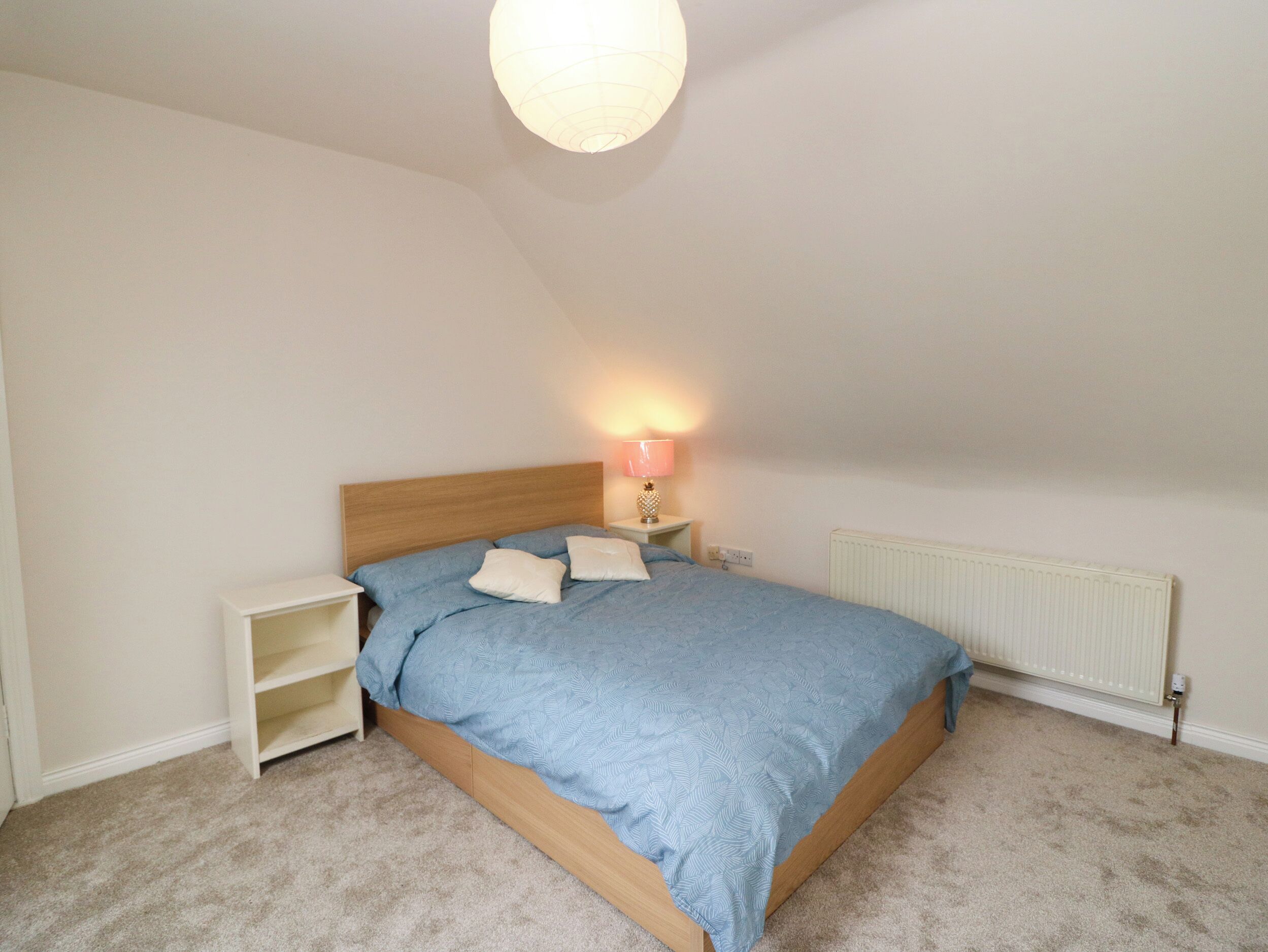 Un dormitorio con techo inclinado, una cama con cabecera de madera, una mesita de noche con lámpara y un radiador.