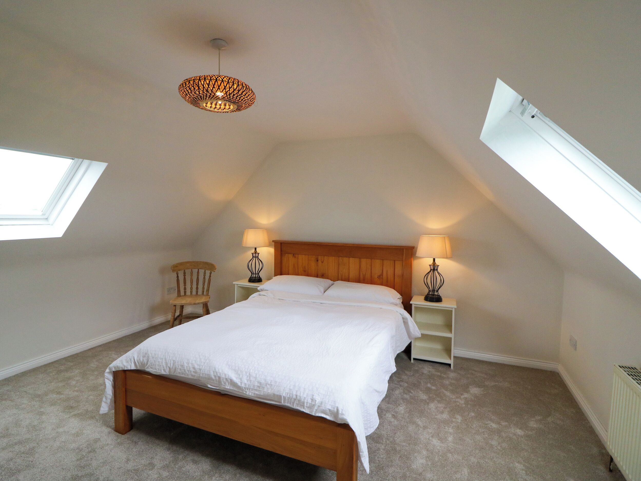 A bedroom with a sloped ceiling, a wooden bed, two bedside tables with lamps, a chair, and a skylight.