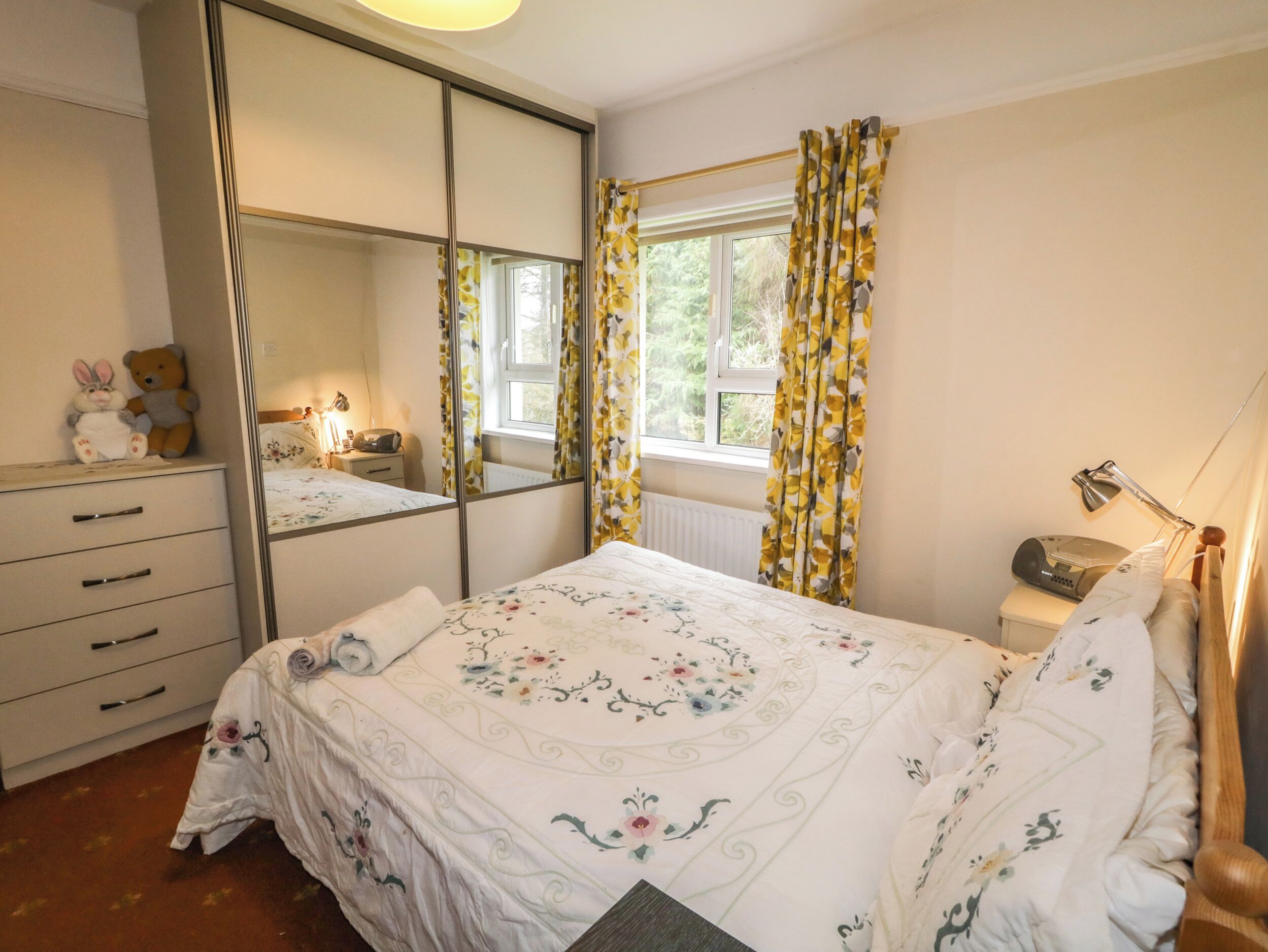 Un dormitorio con cama, cómoda, espejo, ventana con cortinas, lámpara de noche y un osito de peluche sobre la cómoda.