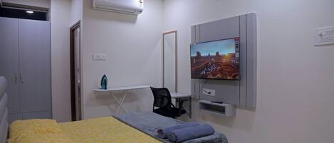 A bedroom with a bed, a desk, a chair, a television, and an air conditioning unit.
