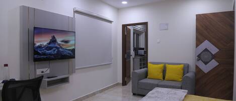 Habitación de hotel moderna con un televisor de pantalla plana en la pared, un sofá gris con cojines amarillos, una cama con colcha estampada en amarillo y blanco, y un escritorio con una silla.