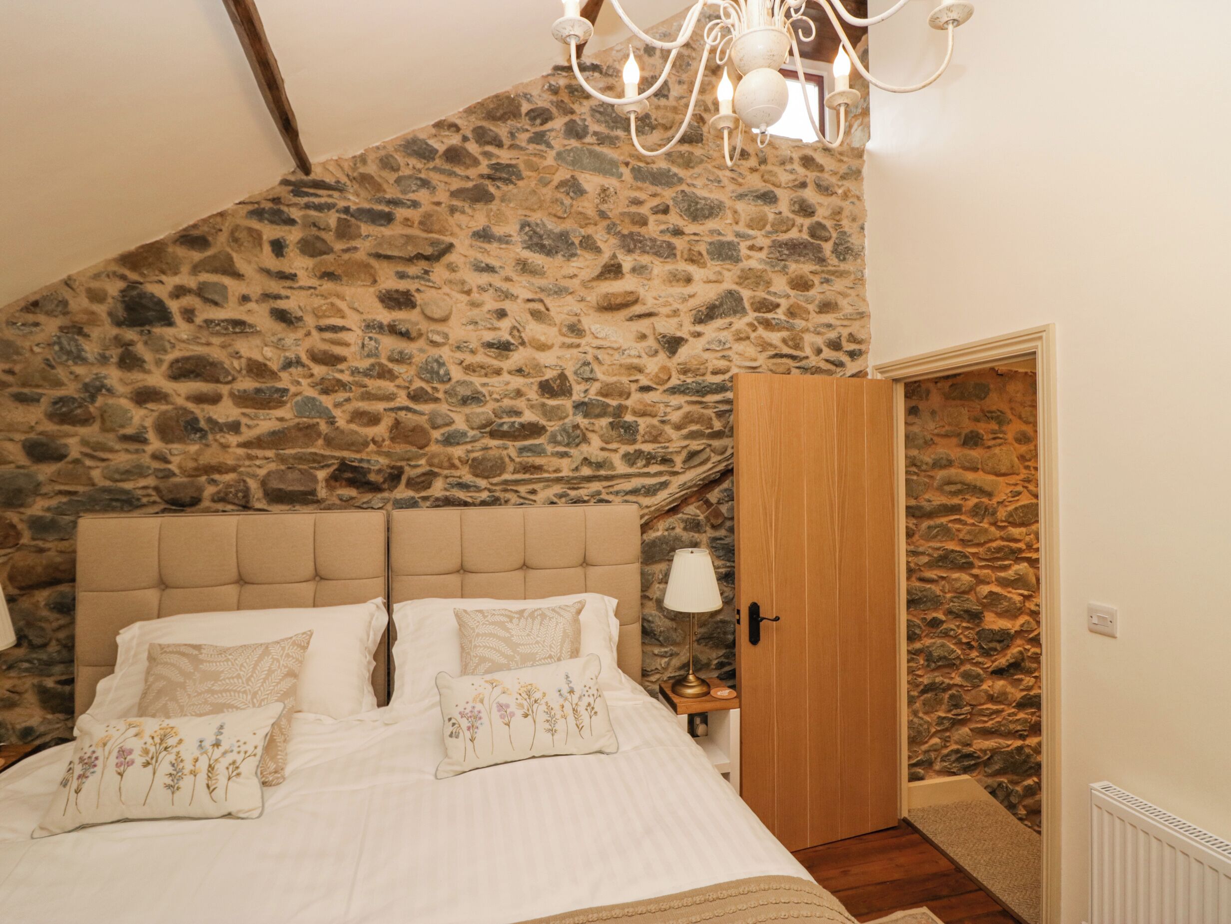 Une chambre avec un mur en pierre, une porte en bois et un lit agrémenté d’oreillers à motifs floraux.