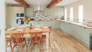 A spacious kitchen with wooden beams, a central island with chairs, and a dining table set for six. - The Hay Barn (Thirsk)