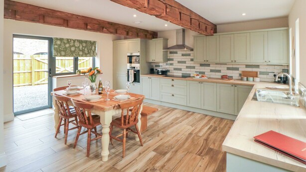 A modern kitchen with wooden beams, a dining area with a wooden table and chairs, and light-colored cabinets. - The Hay Barn (Thirsk)