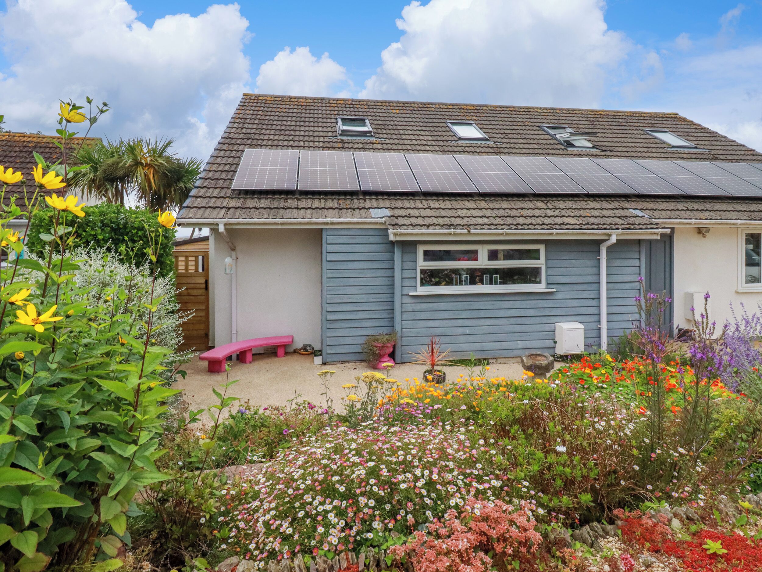 A house with solar panels, a garden with various flowers, and a pink bench.