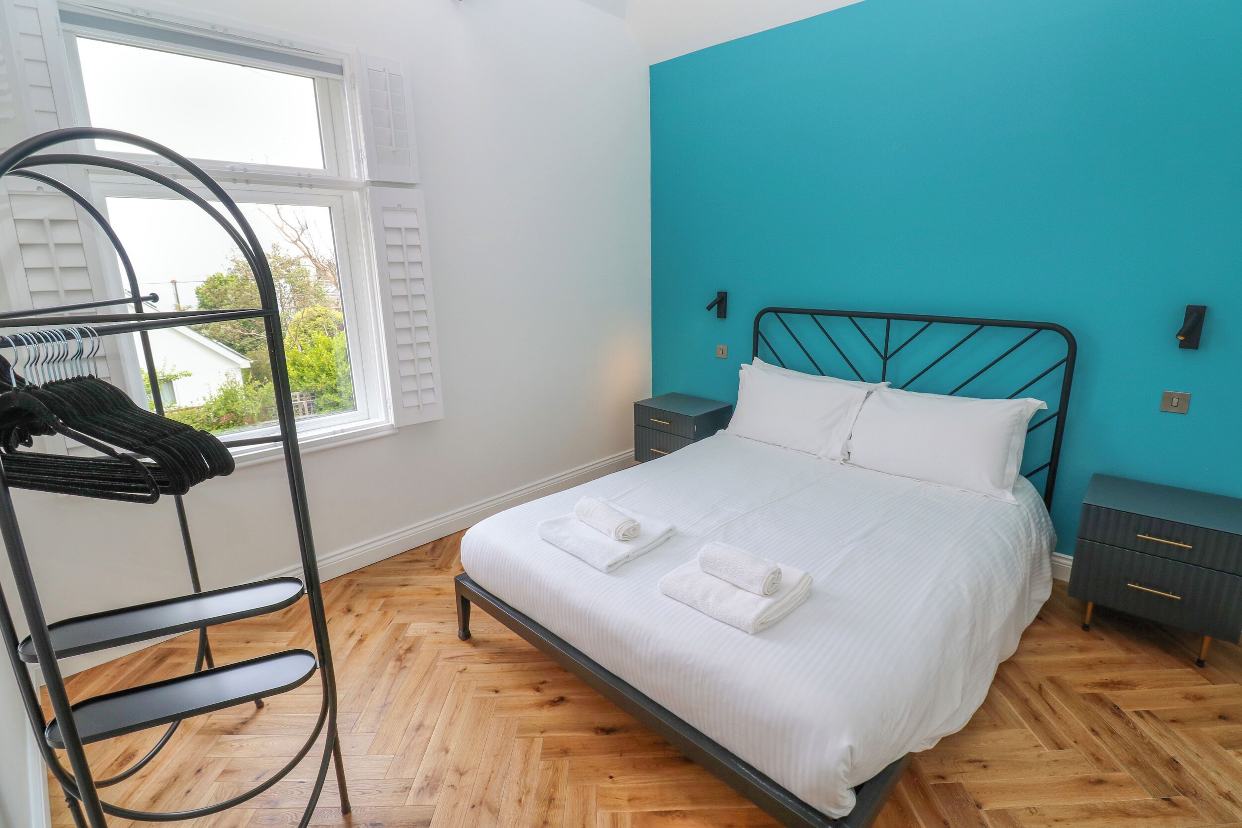 Een net opgemaakt bed met witte lakens en handdoeken, een metalen rek met kleding, een wand in een teal kleur en een houten vloer.