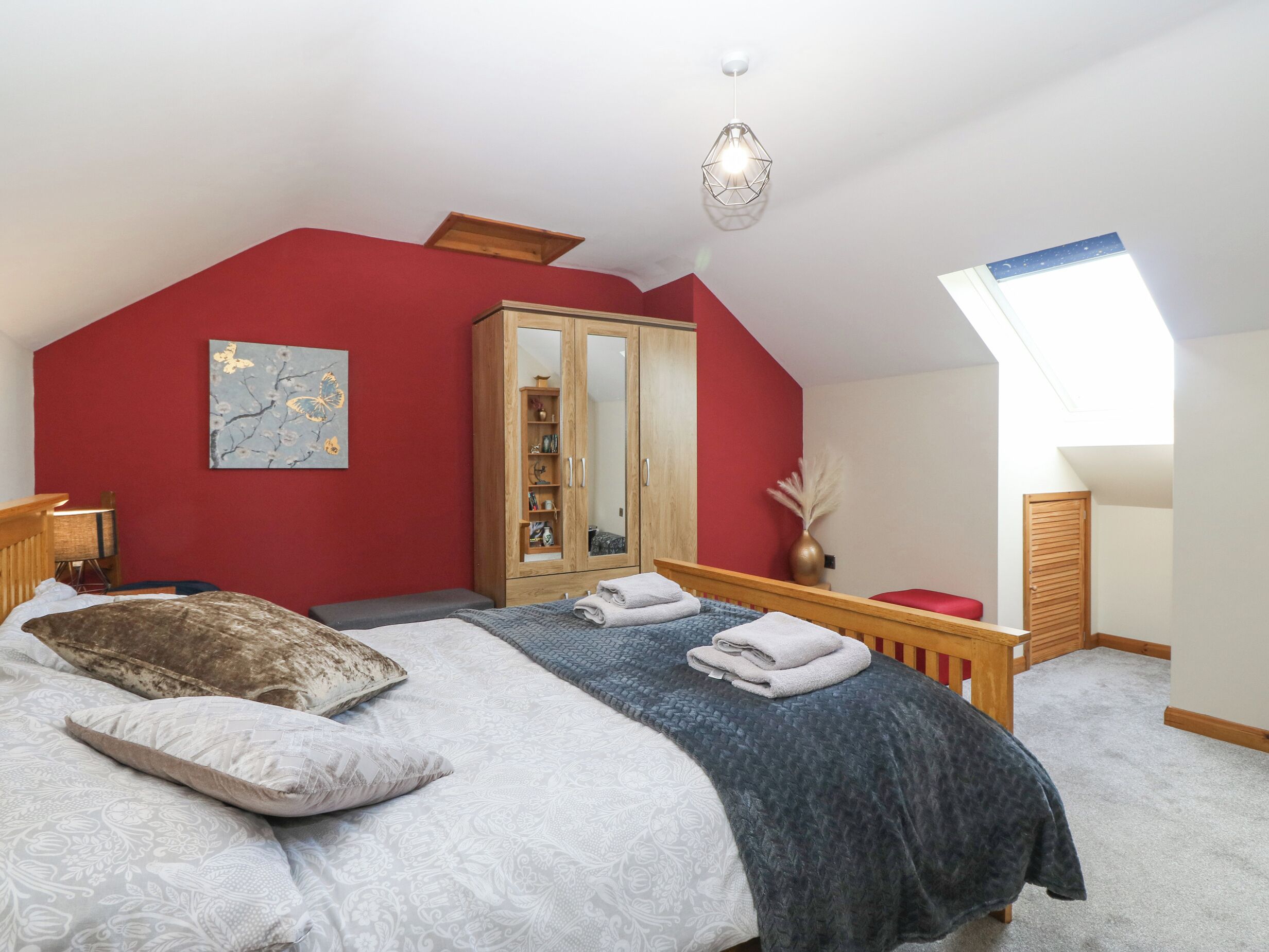 Een slaapkamer met schuine wand, een groot bed met veel kussens, een houten garderobe, een nachtkastje en een dakraam.