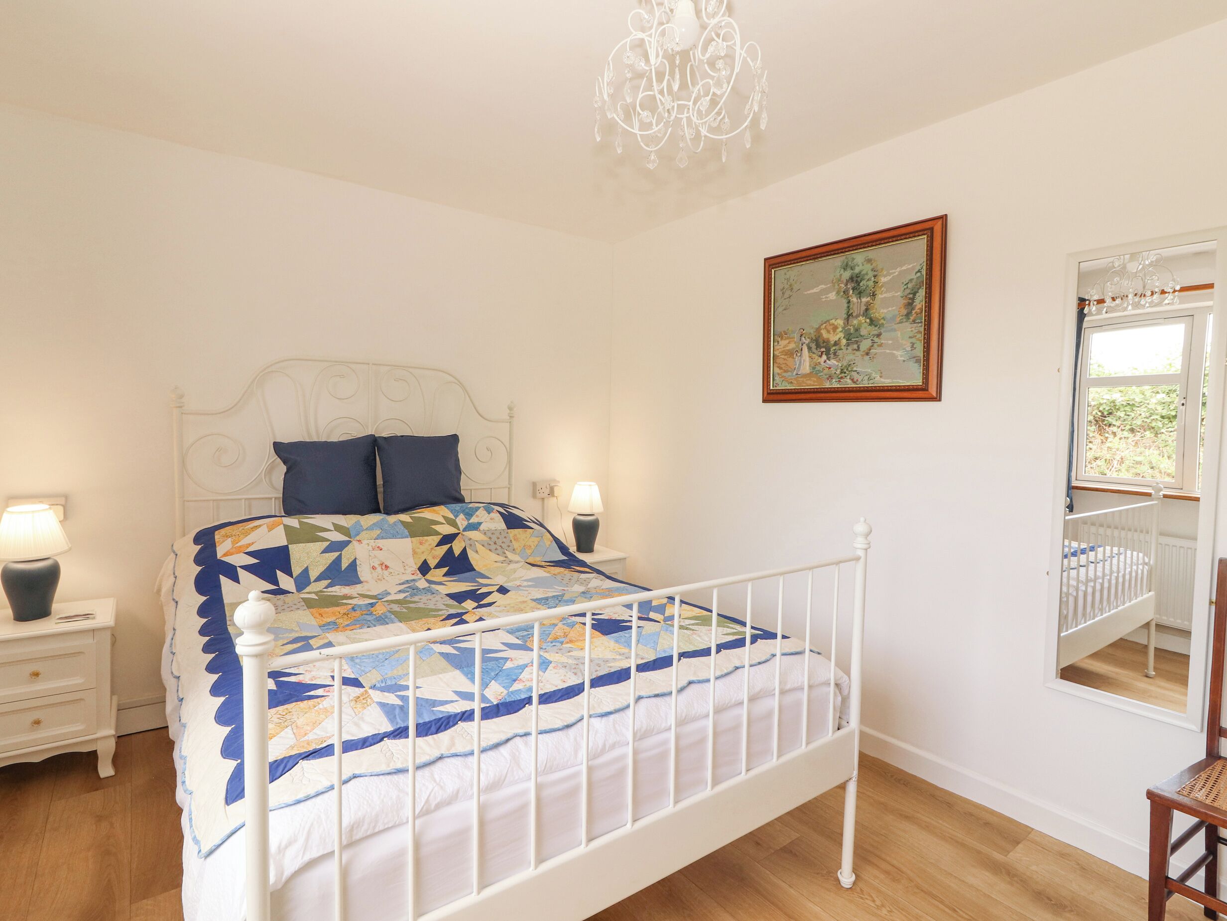 A bedroom with a white metal bed frame, a colorful quilt, two bedside lamps, a painting on the wall, and a window with a view of greenery.