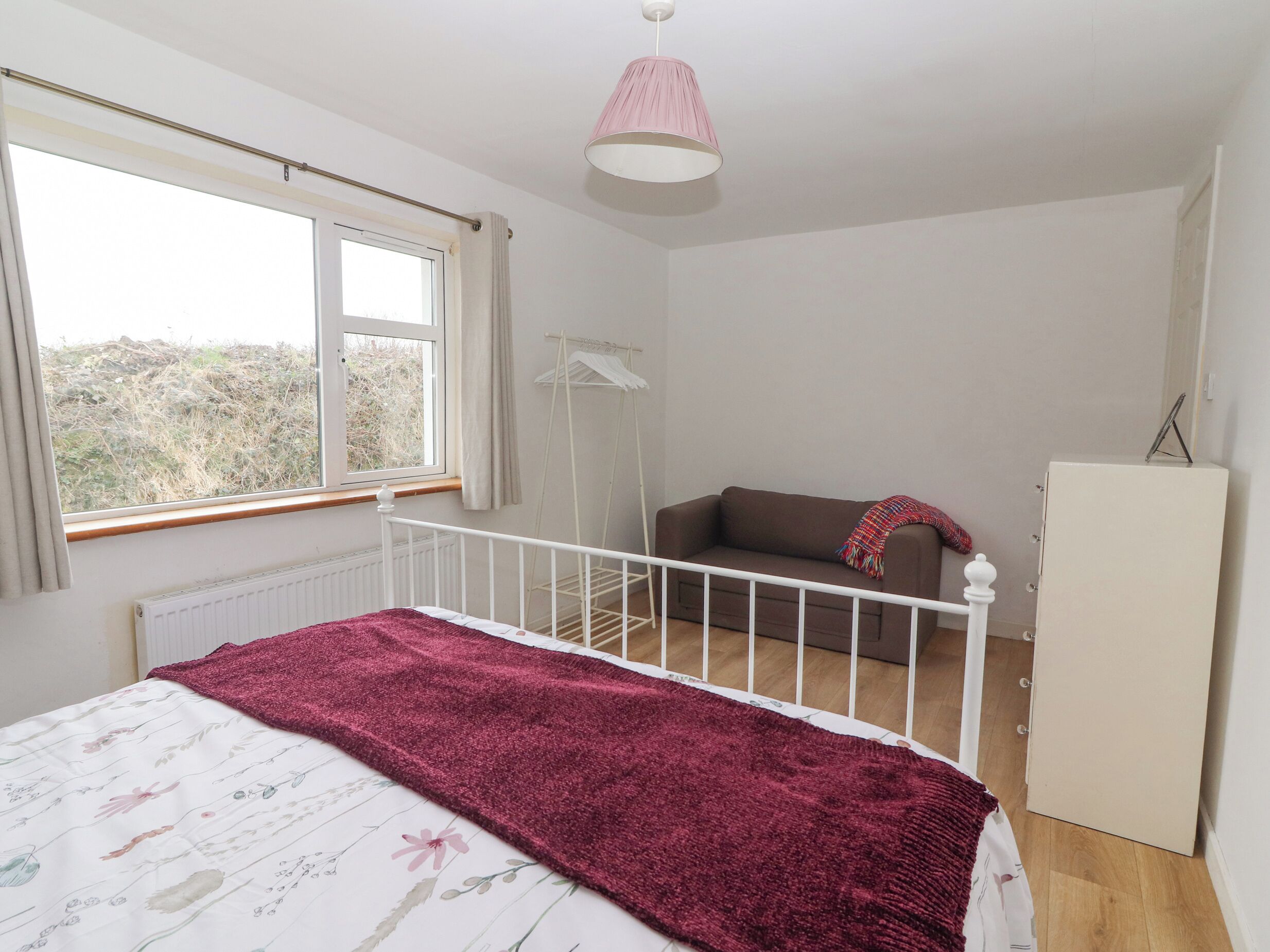 Un dormitorio con cama, ventana, radiador, armario blanco y lámpara colgante.