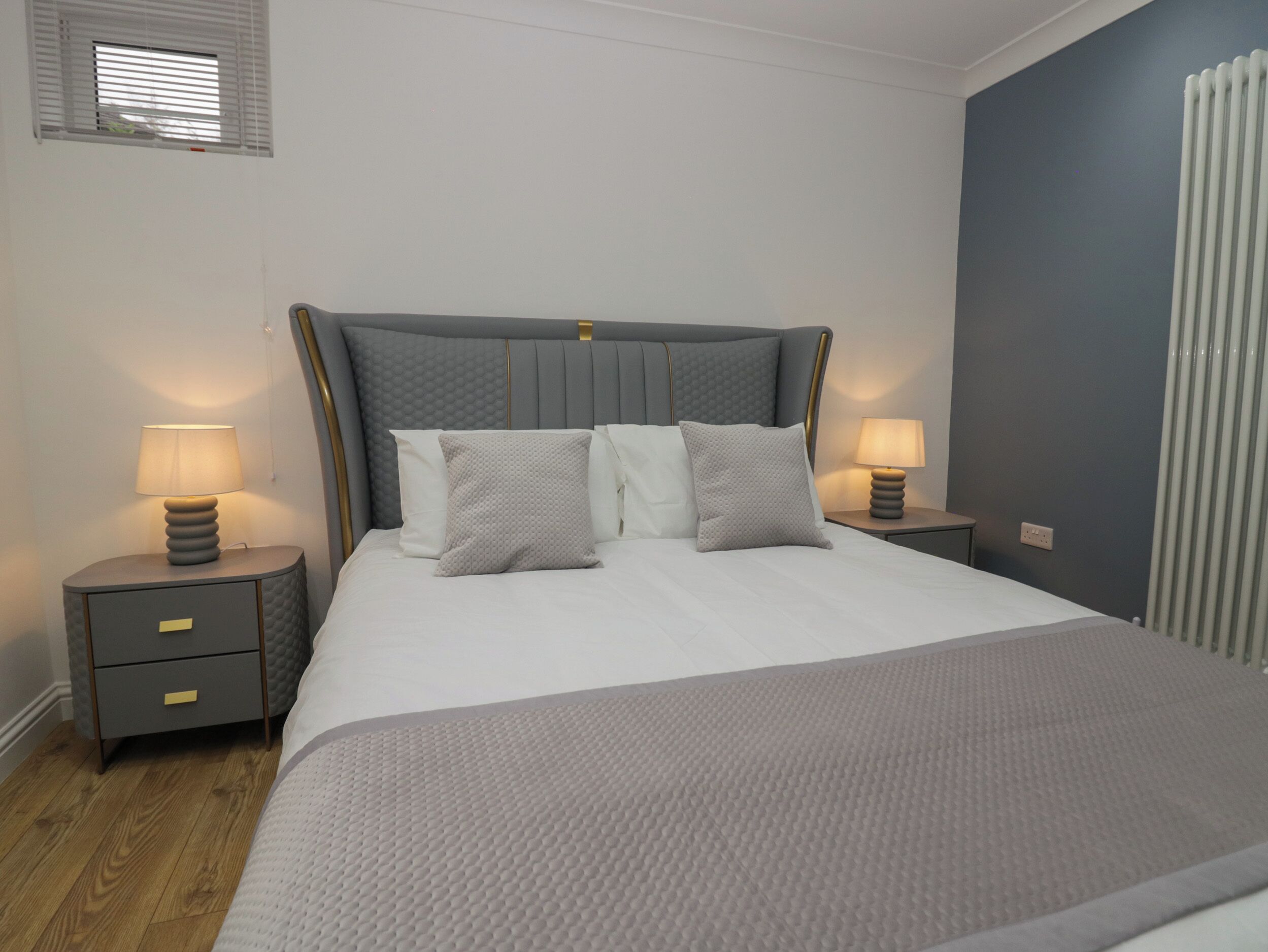 A bedroom with a large bed, two bedside tables with lamps, a window with blinds, and a radiator.
