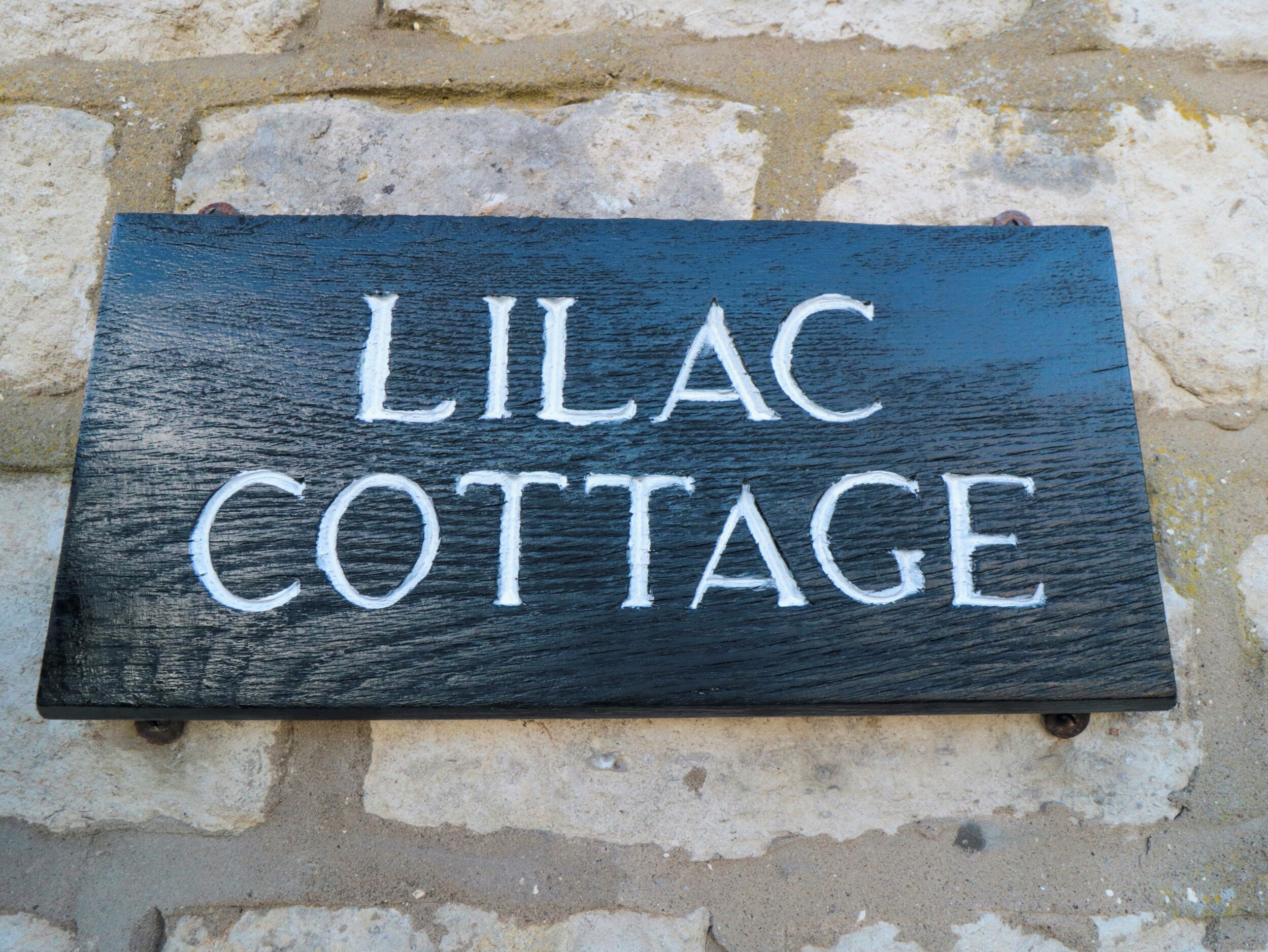 En svart skylt med vit text som visar orden "LILAC COTTAGE", monterad på en stenmur.