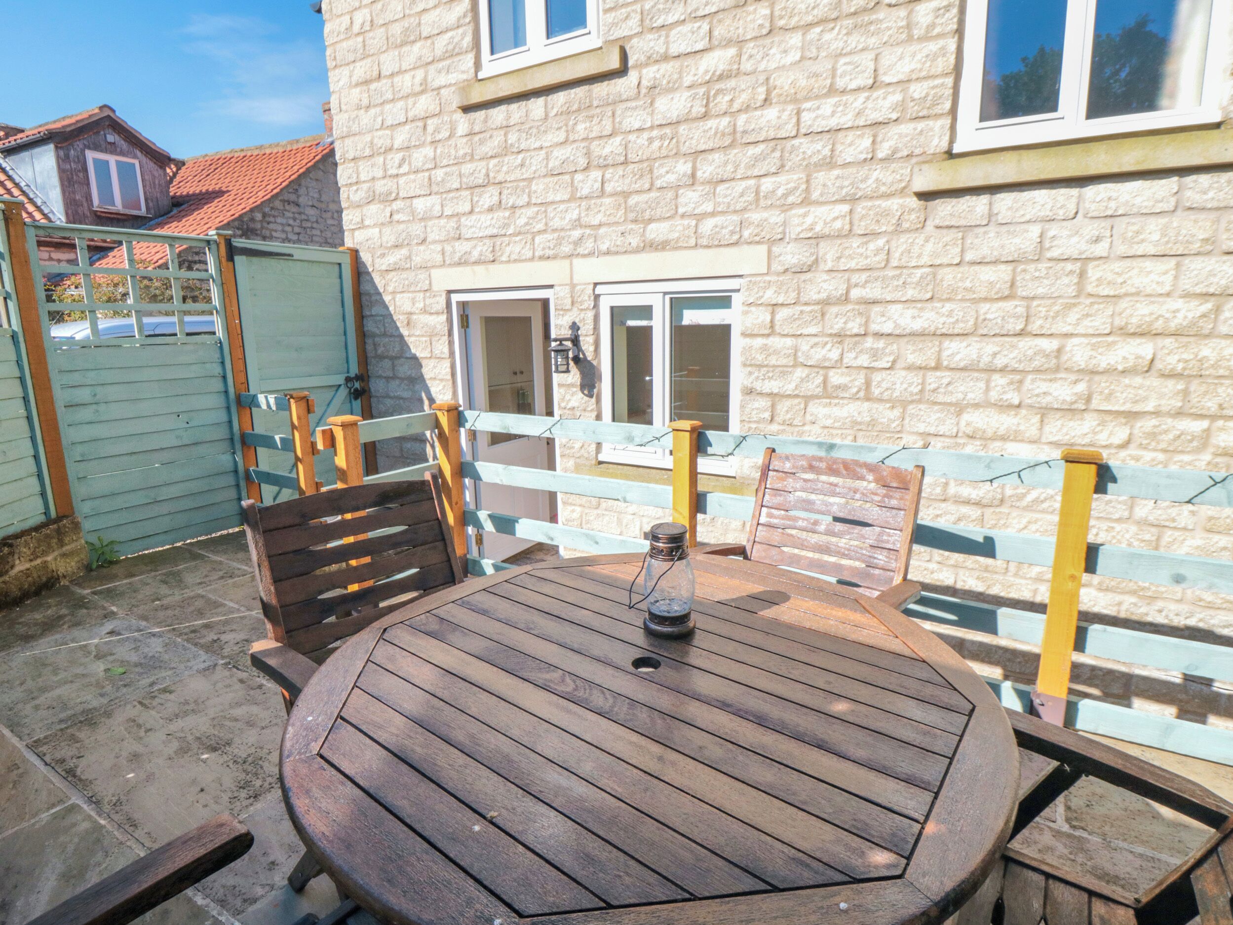A round wooden table with a bottle and menu, surrounded by wooden chairs, against a stone building with a window and door.