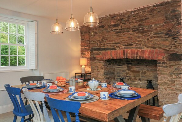 A dining area with a wooden table set for four, surrounded by blue chairs, with a stone wall and a fireplace in the background.