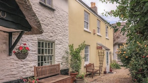 A quaint courtyard with a wooden bench, potted plants, and a thatched roof structure. - Ellenor (Kingsbridge)