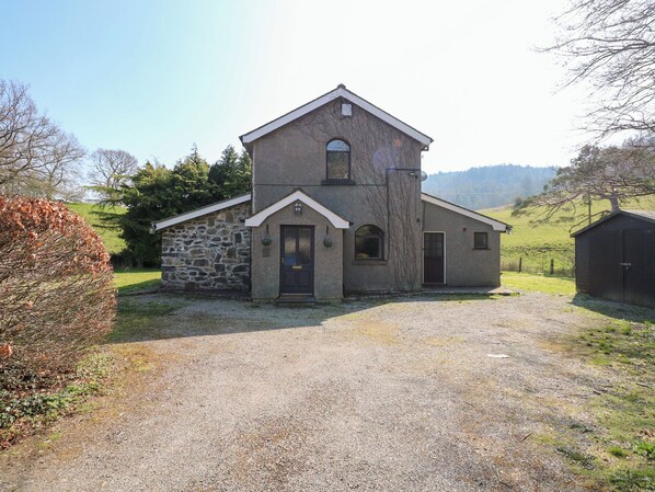 A two-story stone building with a small attached structure, a gravel driveway, and a rural landscape with trees and hills in the background. - Ysgubor Wen (Trefriw)