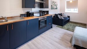 A modern kitchen with dark cabinets, a wooden countertop, and a built-in oven. There is a dining area with a dark sofa and a patterned rug.