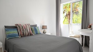A neatly made bed with a gray bedspread and multiple pillows, a bedside table with a lamp, a small white table with a chair, and a window with curtains.