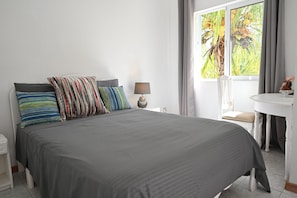 A neatly made bed with a gray bedspread and multiple pillows, a bedside table with a lamp, a small white table with a chair, and a window with curtains.